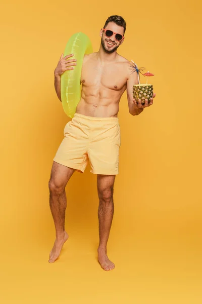 全景尽收眼底 看到戴着太阳镜 拿着泳圈的快乐肌肉男人 还有一半的菠萝和黄色背景的新鲜鸡尾酒 — 图库照片