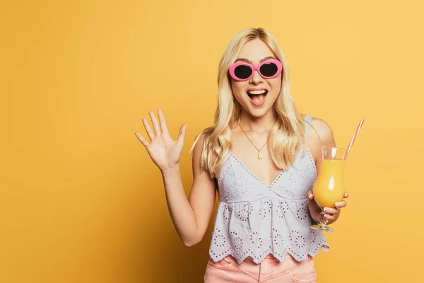 黄色い背景にオレンジジュースのグラスを持ちながら手を振って陽気な女の子 — ストック写真