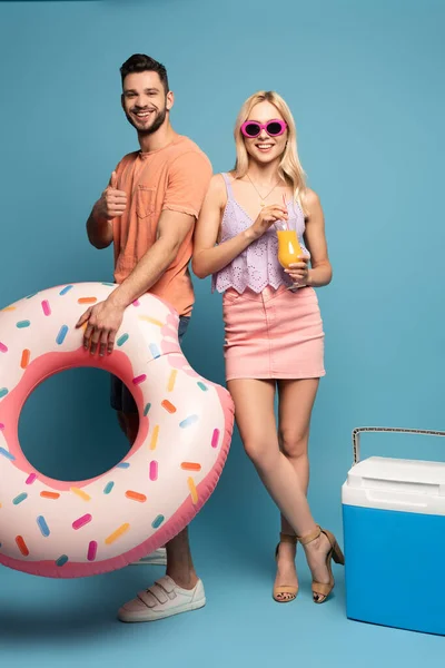 happy man with swim ring showing thumb up, and smiling girl holding glass of orange juice near portable fridge on blue background