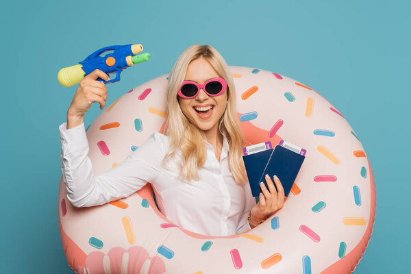 cheerful businesswoman in sunglasses and swim ring holding passports, air tickets and water gun isolated on blue