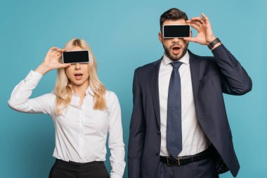 Şaşırtıcı iş adamı ve iş kadını mavi arka planda boş ekranlı akıllı telefonlarla gözlerini kapatıyorlar.