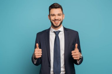 Mutlu iş adamı mavi arka plandaki kameraya bakarken başparmağını kaldırıyor.