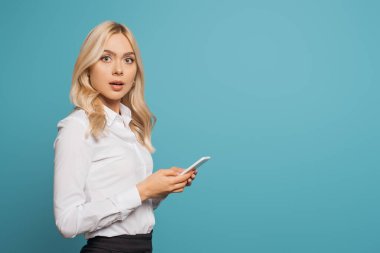 Şok olmuş sarışın iş kadını kameraya bakarken mavi renkli akıllı telefon kullanıyor.