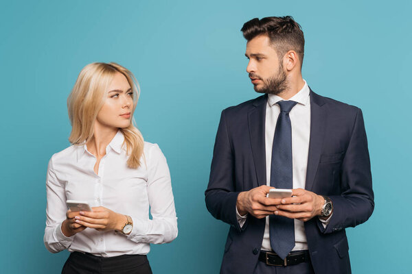 young businesswoman and businessman looking at each other while holding smartphones on blue background