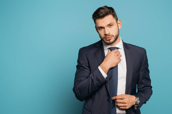 confident, handsome businessman touching tie while looking at camera on blue background