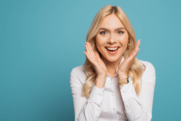 excited businesswoman holding hands near face while looking at camera on blue background
