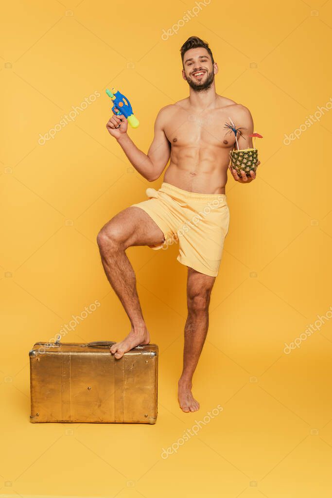 Happy shirtless man holding water gun cocktail in pineapple while stepping on vintage suitcase on yellow background