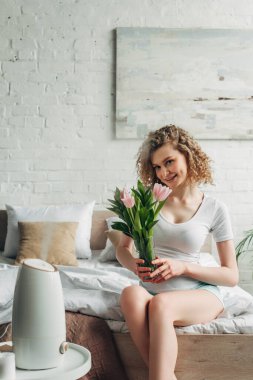 happy girl holding tulips while sitting in bedroom with air purifier  clipart