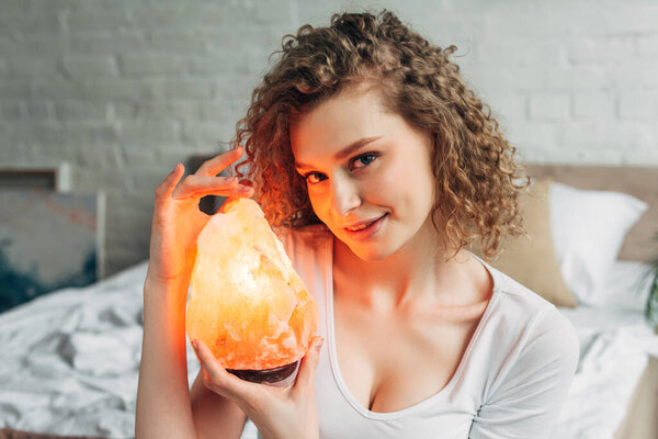 улыбающаяся кудрявая девушка с гималайской соляной лампой в спальне
