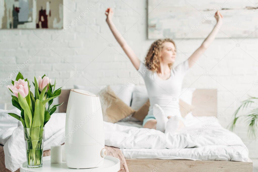 selective focus of happy girl stretching in bedroom with air purifier and tulips 