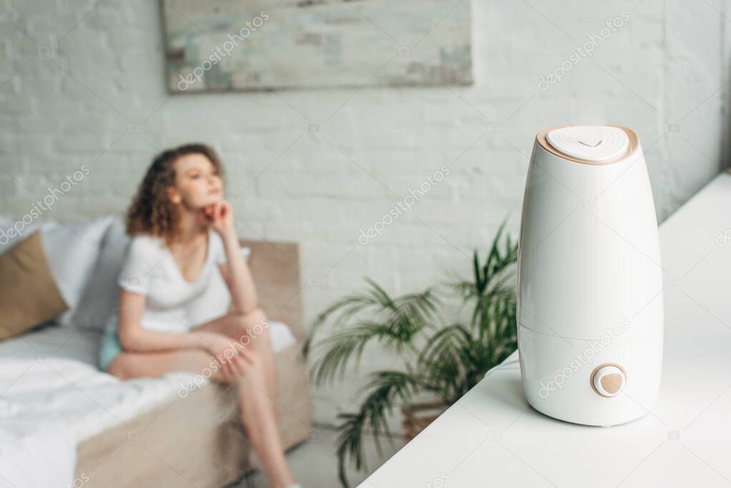 selective focus of attractive girl sitting on bed with air purifier on windowsill