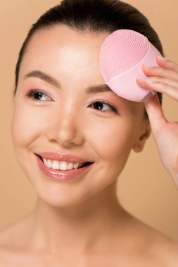 Çekici gülümseyen Asyalı kız pembe silikon temizleyici yüz fırçası kullanıyor.