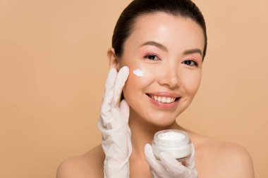 attractive cheerful nude asian girl in latex gloves applying face cream isolated on beige clipart