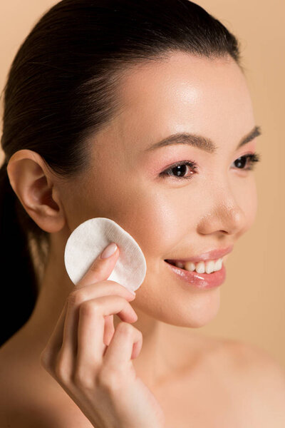 smiling asian girl removing makeup from face with cotton pad isolated on beige