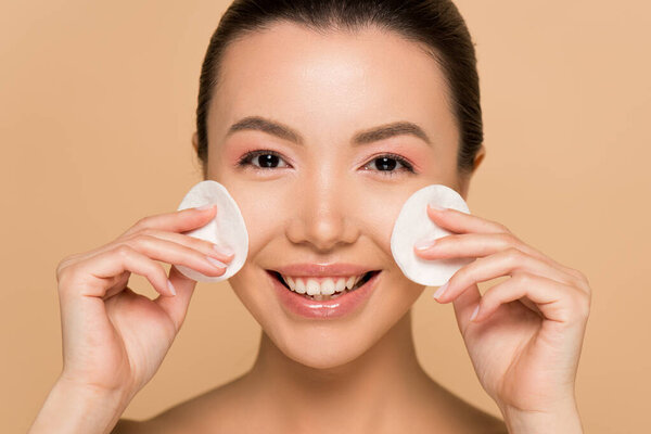 smiling asian woman removing makeup from face with cotton pads isolated on beige