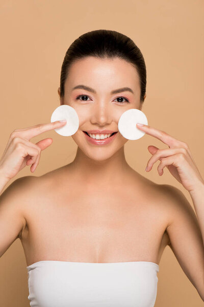 cheerful asian girl removing makeup from face with cotton pads on beige