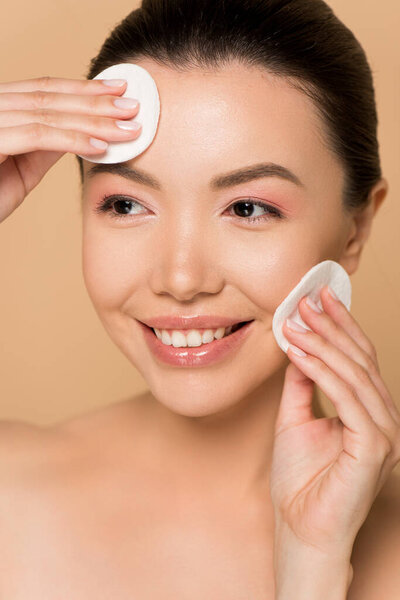 beautiful happy asian woman removing makeup from face with cotton pads isolated on beige