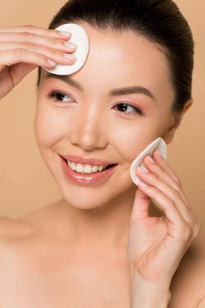 attractive smiling nude asian woman removing makeup from face with cotton pads isolated on beige