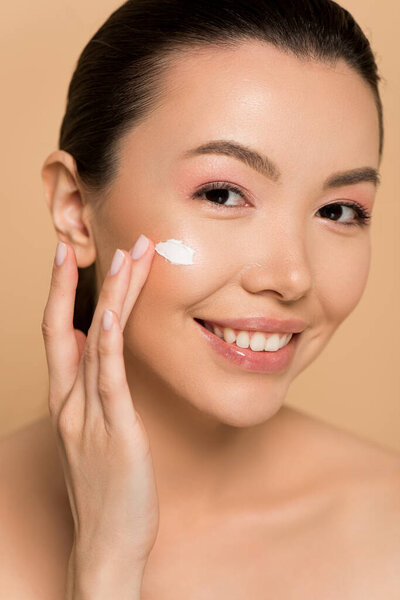 attractive naked asian girl applying cosmetic cream on face isolated on beige