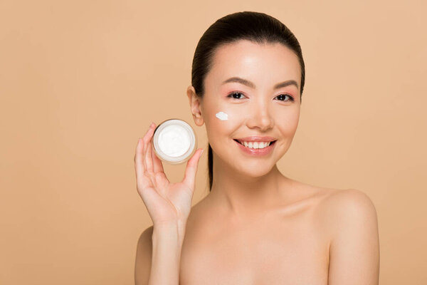 beautiful smiling naked asian girl holding glass container with cosmetic cream isolated on beige