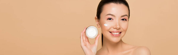 panoramic shot of beautiful naked asian girl holding glass container with cosmetic cream isolated on beige