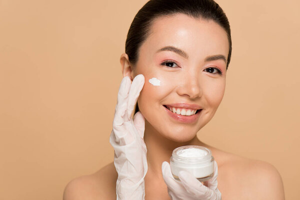 attractive cheerful nude asian girl in latex gloves applying face cream isolated on beige