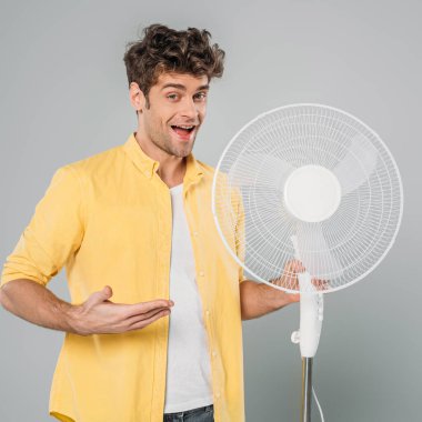Heyecanlı ve ağzı açık bir adam kameraya bakıyor ve gri renkte izole edilmiş elektrikli vantilatörü işaret ediyor.