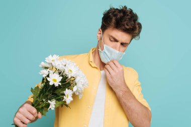Tıbbi maskeli bir adam çiçekleri tutuyor ve mavi üzerine uzanmış elini uzatıyor.