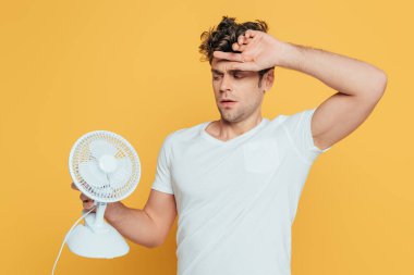 Confused man with raised hand looking at desk fan isolated on yellow clipart