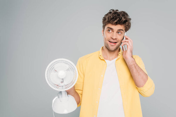 Front view of man smiling, looking away, holding desk fan and talking on smartphone isolated on grey