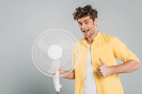 Man with open mouth holding electric fan, looking at camera and showing like sign isolated on grey