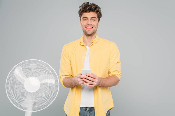 Вид человека, улыбающегося, смотрящего на камеру и держащего смартфон рядом с электрическим вентилятором, изолированным на сером фоне
