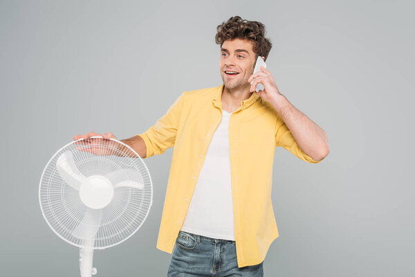 Мужчина улыбается и разговаривает на смартфоне рядом с электрическим вентилятором, изолированным на сером
