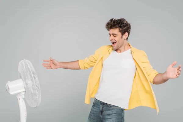 Excited man with open arms enjoying electric fan isolated on grey