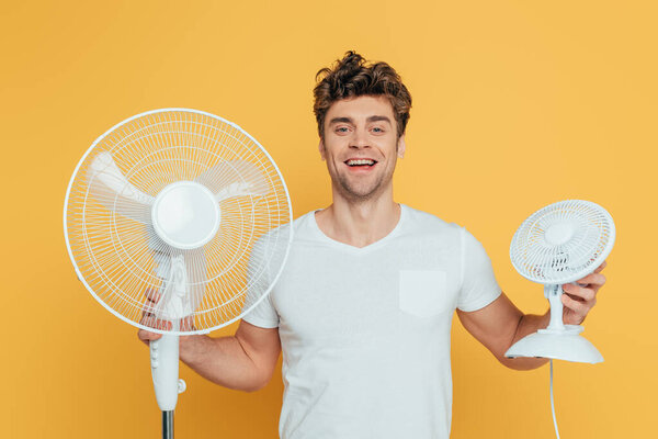Front view of man holding electric and desk fans, smiling and looking at camera isolated on yellow