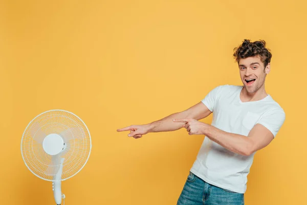 興奮した男はカメラを見て 黄色に隔離された電気ファンを指して — ストック写真