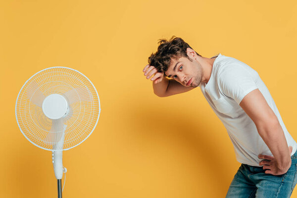 Man with hand on hip looking at camera and leaning near electric fan on yellow