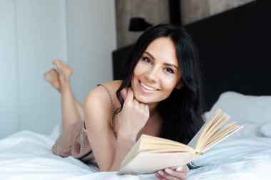 Evdeki yatakta kitap okuyan güzel, gülümseyen kız.
