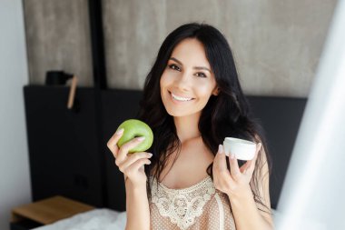 Gülümseyen kadın yatak odasında elma ve plastik kapla kozmetik krem taşıyor.