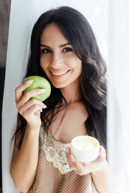 Yeşil elma tutan pozitif kadın ve yüz kremi sürmüş plastik kap 