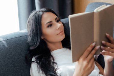 Evdeki kanepede kitap okuyan güzel, gülümseyen kız.