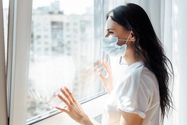 больная молодая женщина в медицинской маске смотрит в окно на самоизоляцию
