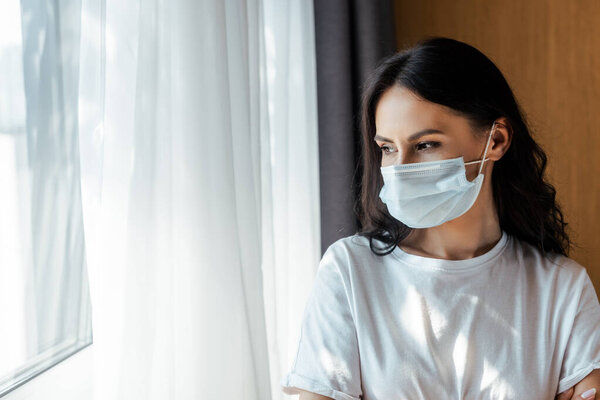 attractive ill woman in medical mask looking through window on self isolation