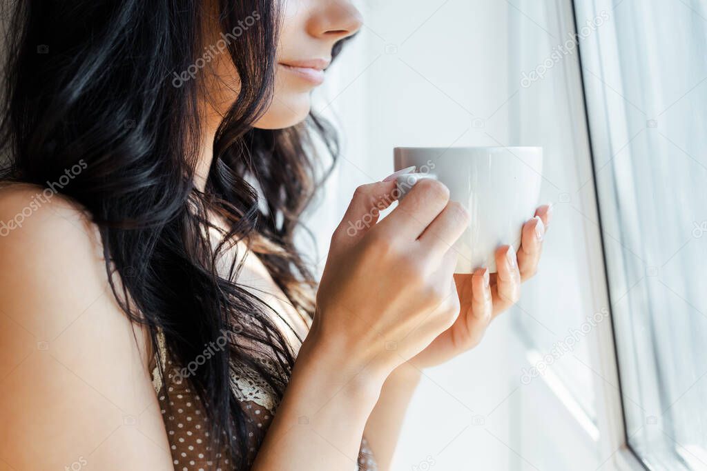 cropped view of girl holding cup of coffee near window 