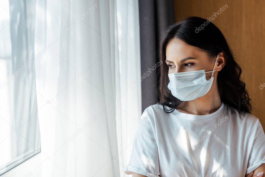 attractive ill woman in medical mask looking through window on self isolation