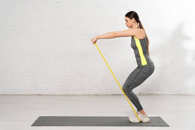 Beyaz tuğla duvarın yanında spor giyinen bir kadının spor giyinme bandıyla spor yaparken çekilmiş yan görüntüsü. 