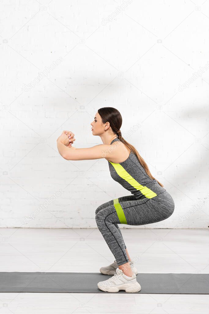 side view of athletic girl doing squat while exercising with resistance band on fitness mat
