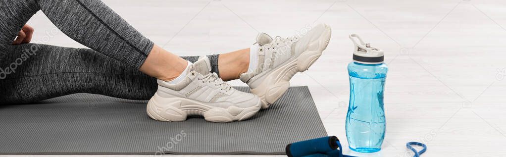 panoramic orientation of sportswoman sitting on fitness mat near sports bottle with water and skipping rope 