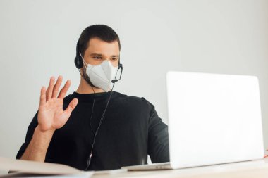 Tıbbi maskeli ve kulaklıklı operatörün seçici odak noktası evde video görüşmesi yaparken el sallaması