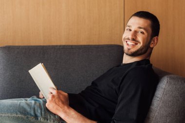 Gülümseyen ve sakallı bir adam oturma odasında kitap tutuyor. 